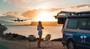 Woman standing next to campervan overlooking the sea and a plane starting during sunset.