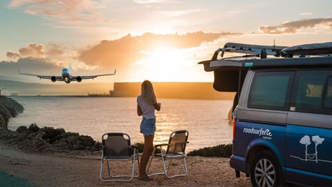 Woman standing next to campervan overlooking the sea and a plane starting during sunset.