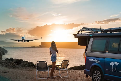 Woman standing next to campervan overlooking the sea and a plane starting during sunset.