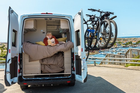 Woman is lying on a bed inside the back of a camper van, reading a book, with two bicycles mounted on a rear rack and a scenic harbor view in the background.