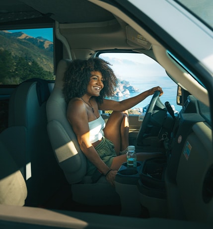 Smiling woman with curly hair sitting in the driver’s seat of a motorhome, enjoying a scenic coastal view through the window