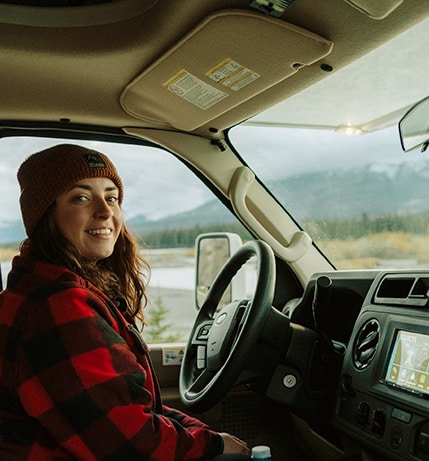 Smiling woman wearing a red plaid jacket and a beanie, sitting in the driver’s seat of a motorhome with a scenic mountainous landscape visible through the windshield