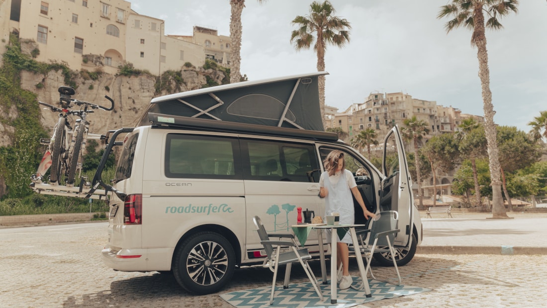 woman in a summerdress, campingtable in front of a campervan, city and palm trees in the background