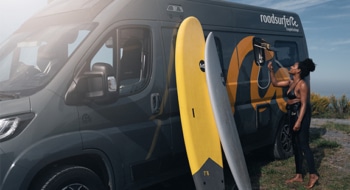 Young woman using an outdoor shower attached to a roadsurfer campervan parked by the coast, with two surfboards leaning against the vehicle.