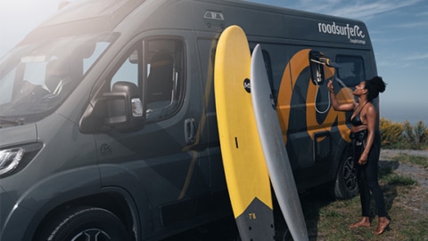 Young woman using an outdoor shower attached to a roadsurfer campervan parked by the coast, with two surfboards leaning against the vehicle.