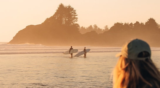 Woman wearing a cap watching two surfers walking on a beach during sunset.