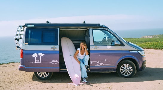 Woman with surfboard sitting in a roadsurfer camper van in the sunshine