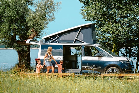 A mother walks hand in hand with her two children along the coast in front of a blue VW campervan with an open pop-up roof, enjoying a sunny day by the seaside.