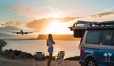 Woman standing next to campervan overlooking the sea and a plane starting during sunset.