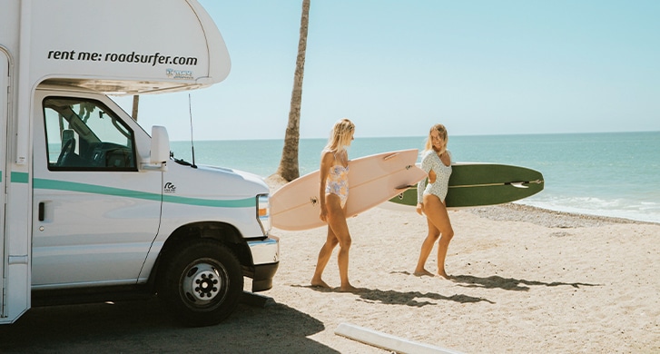 Women On The Beach And Next To Rv Carrying Surfboards US