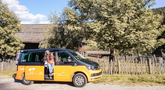 Smiling woman in a black and yellow campervan in the countryside, wooden fence and huge green tree in the background